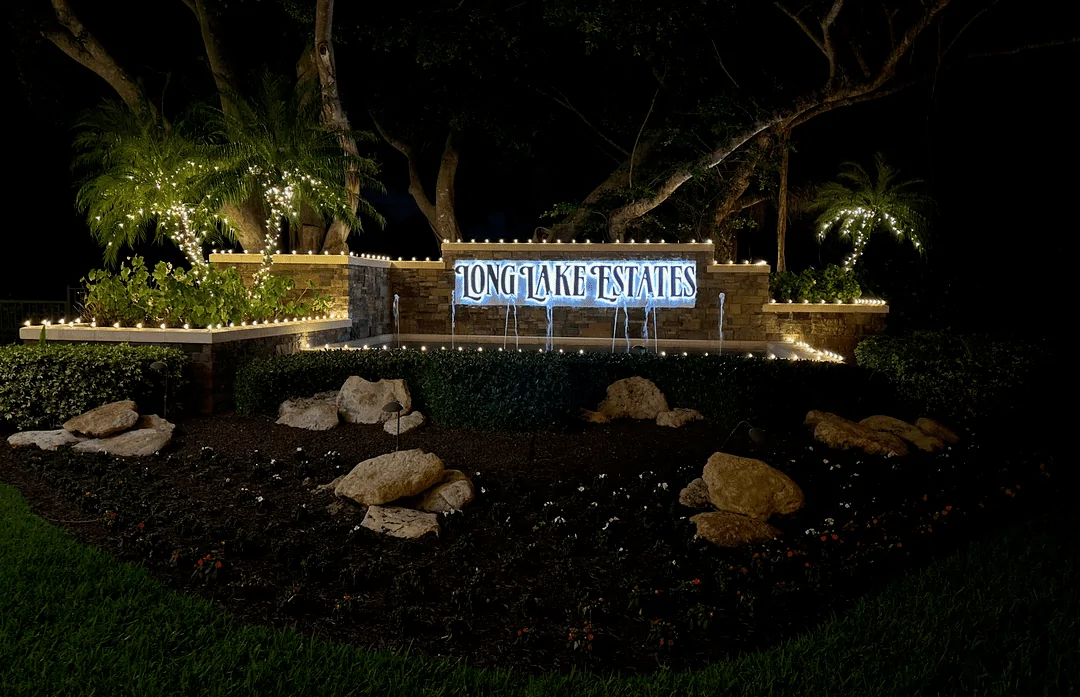 Long Lake Estates stone monument sign with backlit lettering, water fountains, and lit palms at night