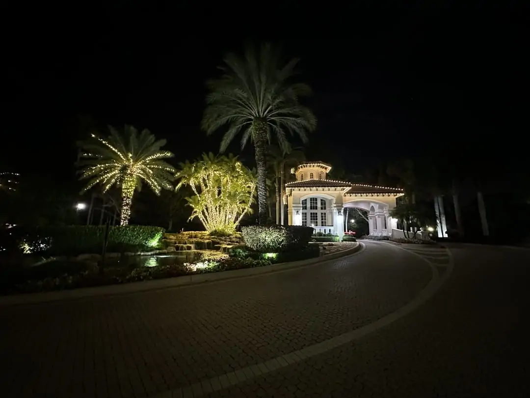 Gated community clubhouse with warm white roofline lighting and palms wrapped in lights along a curved paver driveway at night