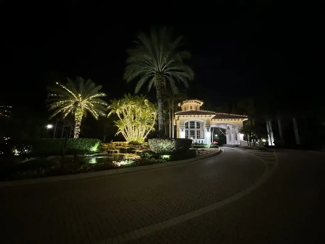 Gated community clubhouse with warm white roofline lighting and palms wrapped in lights along a paver driveway at night